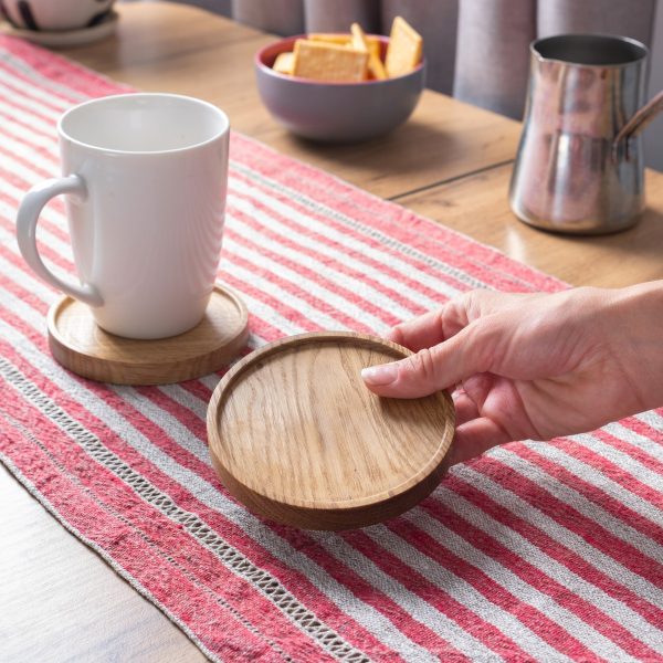 A hand holds a round wooden coaster next to a white mug on a red and white striped table runner.