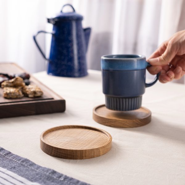 A hand lifts a blue ceramic mug from a round wooden coaster on a table.
